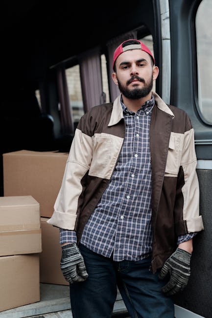A man with a beard and wearing a black cap and white polo shirt stands with arms crossed in front of an open moving van filled with packed cardboard boxes of various sizes. The boxes are stacked insid